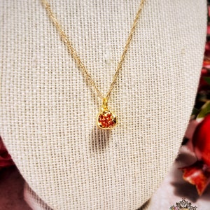 May include: A gold chain necklace with a small red gemstone pendant shaped like a pomegranate. The necklace is displayed on a beige fabric mannequin.