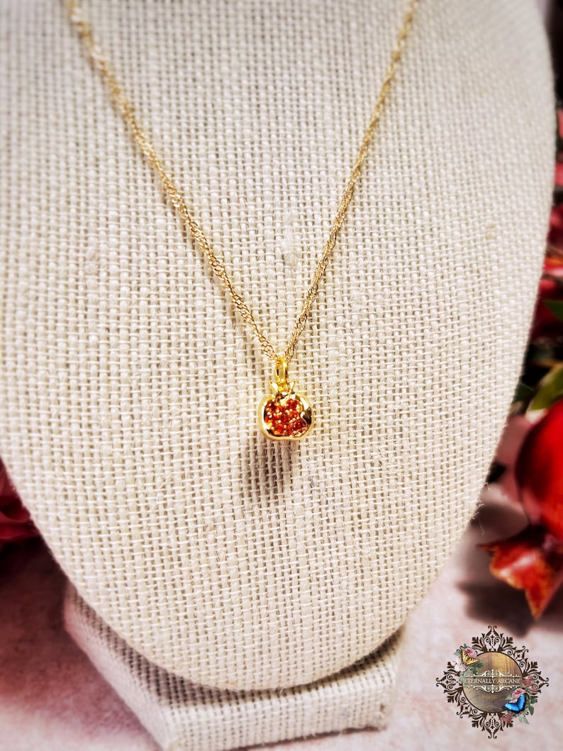 May include: A gold chain necklace with a small red gemstone pendant shaped like a pomegranate. The necklace is displayed on a beige fabric mannequin.