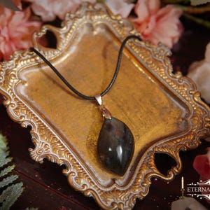 May include: A pendant necklace with a dark, teardrop-shaped stone suspended from a black cord. The pendant is set in a silver-colored metal. The necklace rests on a gold-colored, ornate tray, surrounded by pink roses and greenery.