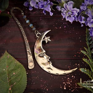 May include: Silver bookmark featuring a crescent moon with a face and a star, connected to a beaded chain. The bookmark has an ornate design and is surrounded by flowers and leaves. The text "Eternally Arcane" is visible.