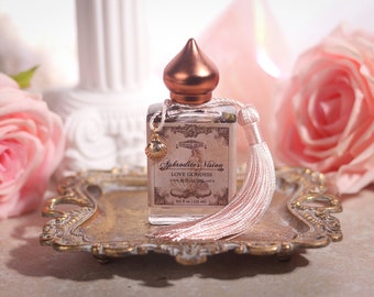 May include: A small glass bottle with a rose gold lid and a pink tassel. The bottle is filled with a clear liquid and has a label that reads "Aphrodite's Vision Love Goddess Love Ritual Oil 0.5 fl oz (15 ml)". The bottle is sitting on a gold tray with two pink roses in the background.