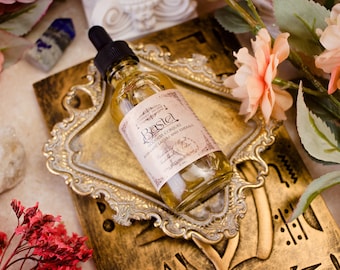 May include: A glass bottle with a gold label that reads "Bastel Golden Copal with Labdanum, Lazuli and Embers" on a gold tray with a floral background.