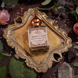 May include: A square glass bottle with a copper-colored cap sits on a gold-colored ornate tray. The bottle has a label that reads "Witch's Ward Spiritual Protection Black Tourmaline". The tray is surrounded by greenery and flowers.