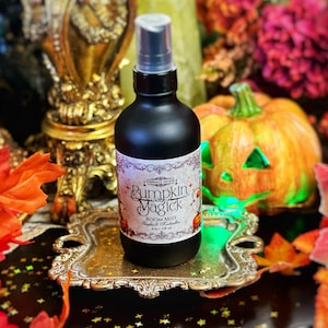 May include: A black bottle of room mist with a white label that says "Pumpkin Jagick Room Mist" with a pumpkin and witch design. The bottle is sitting on a silver tray with fall leaves and a carved pumpkin in the background.