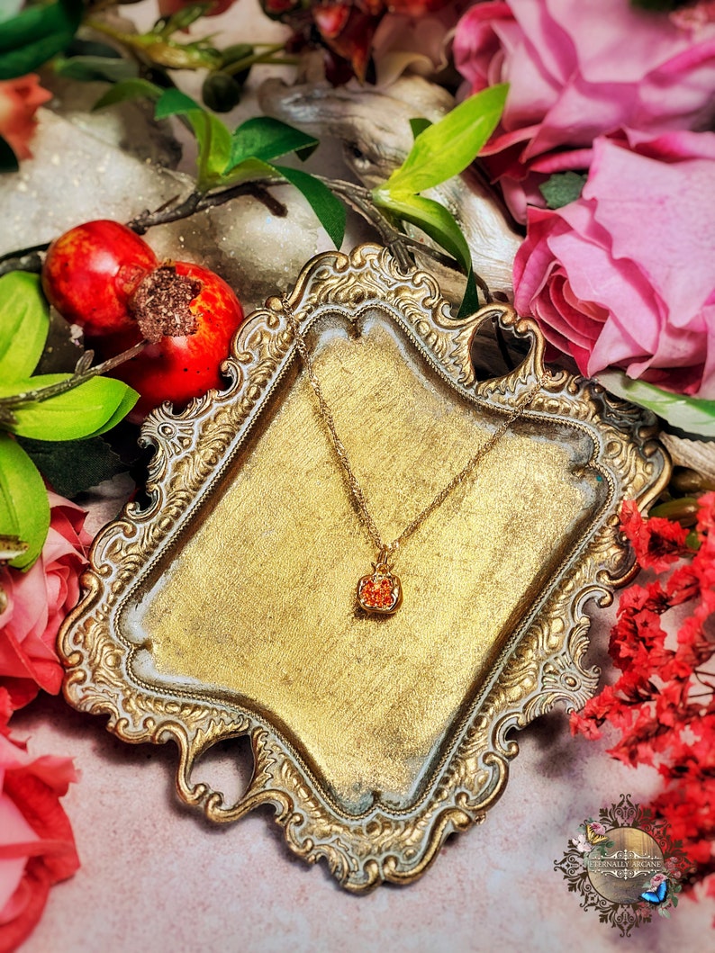 May include: A gold-toned necklace with a small, orange, round pendant on a decorative, ornate, gold-toned tray. The tray is surrounded by pink and red flowers.