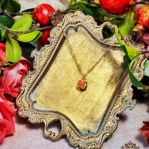 May include: A gold-tone necklace with a small, red, crystal-encrusted pendant shaped like a pomegranate. The necklace is displayed on a decorative, ornate, gold-tone tray.
