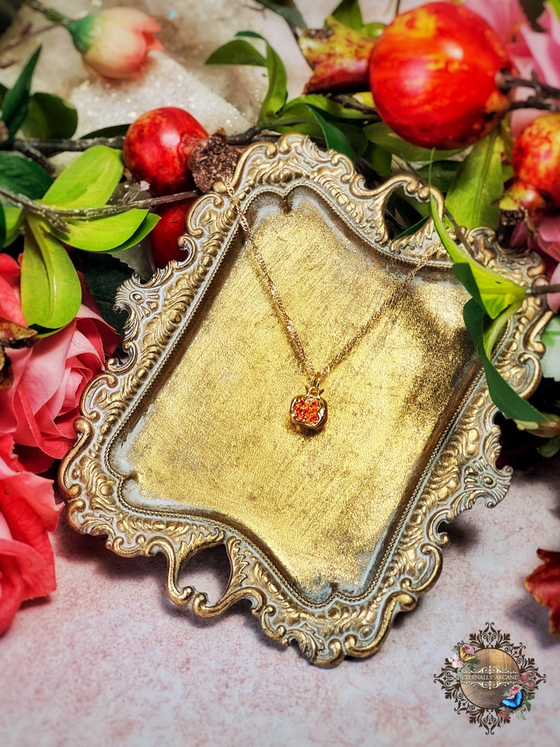 May include: A gold-tone necklace with a small, red, crystal-encrusted pendant shaped like a pomegranate. The necklace is displayed on a decorative, ornate, gold-tone tray.