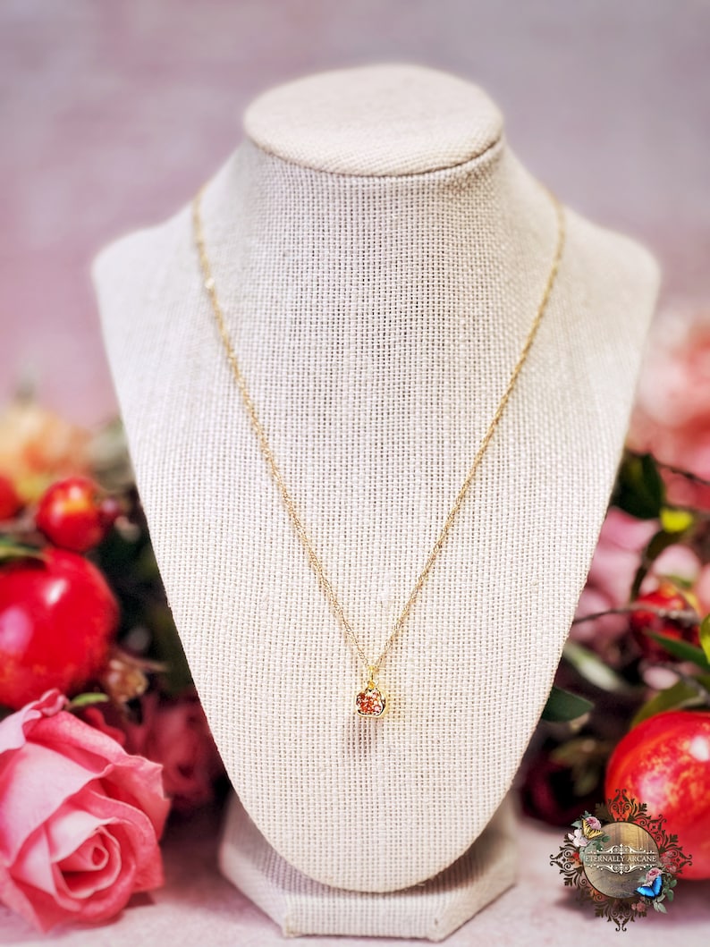 May include: A gold chain necklace with a small, red, sparkly pendant. The necklace is displayed on a fabric-covered mannequin.