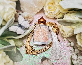 Puede incluir: Un marco decorativo dorado con un borla blanca, que contiene una pequeña botella de vidrio con una tapa dorada y un amuleto de media luna. La botella contiene un líquido transparente y tiene una etiqueta con el rostro de una mujer y la palabra "Oracle". El marco está rodeado de flores blancas y una mariposa blanca.