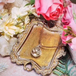 May include: A delicate gold-toned necklace with a round glass pendant containing a dried dandelion. The necklace is displayed on a small, ornate, gold-toned tray with a white background. The tray is surrounded by pink and white flowers.