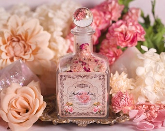 Puede incluir: Una botella de vidrio llena de sales de baño rosas y pétalos de rosa. La etiqueta dice "Ardent Rose" con el texto "Luxury Ritual Bath Salt" y "8 oz / 226 g".