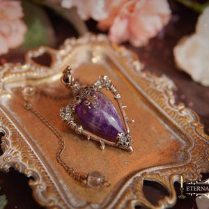 May include: A vintage-style pendulum necklace with a large, polished purple gemstone pendant. The pendant is set in an ornate, antique-gold colored metal frame. The necklace rests on a decorative, gold-toned tray, surrounded by flowers.