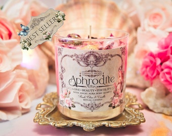 Puede incluir: Vela de vidrio rosa con una etiqueta que dice "Eternally Arcane Aphrodite Love - Beauty - Fertility with Angel Aura Rose Quartz Rose, Citrus & Vanilla Ritual Candle 11 oz". La vela está en una bandeja de metal dorado y rodeada de rosas rosas.