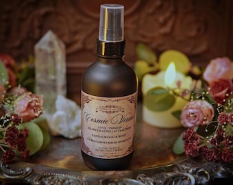 May include: A dark brown spray bottle labeled "Cosmic Venus" with a clear cap, surrounded by flowers, crystals, and a lit candle. The bottle contains a ritual body and sacred space spray.