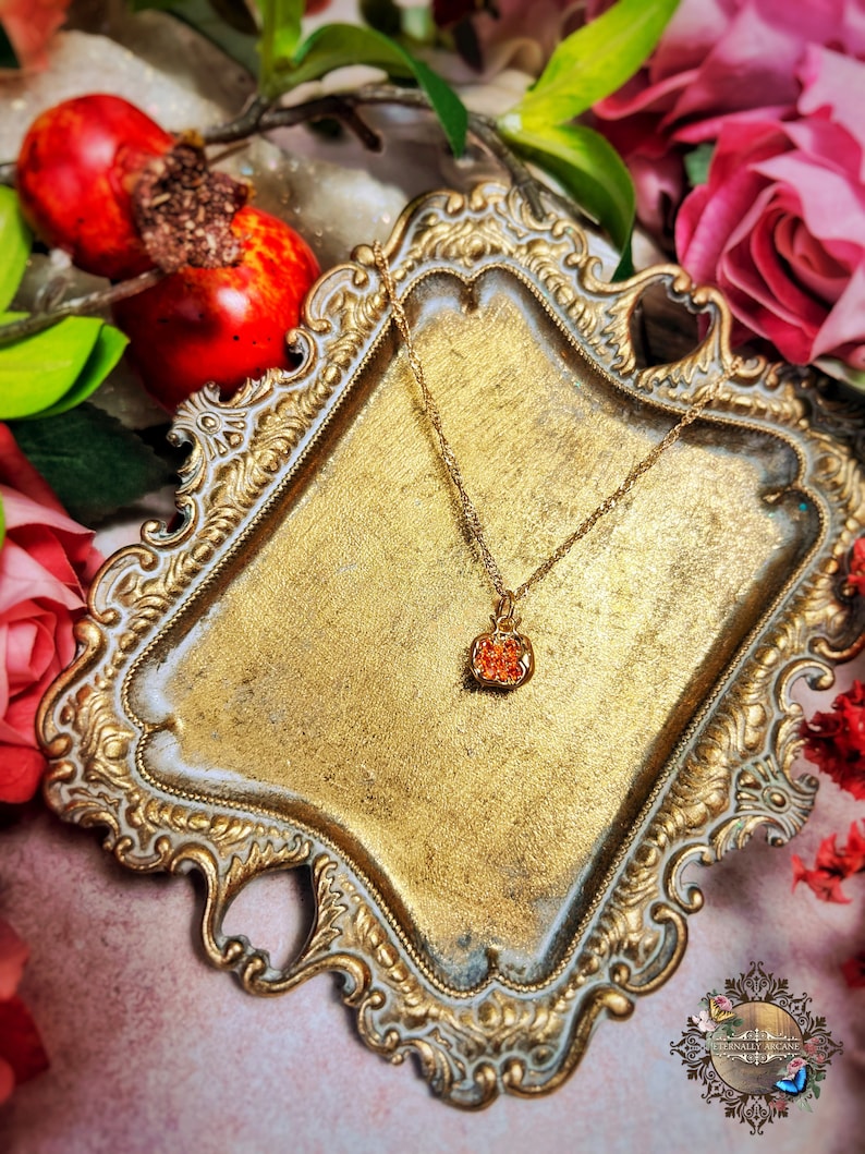 May include: A gold necklace with a small, orange, square pendant on a gold, ornate, decorative tray.