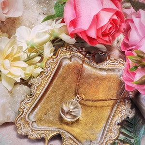 May include: A delicate necklace with a glass pendant containing a dried dandelion. The necklace is displayed on a gold-colored tray with a brown cord.