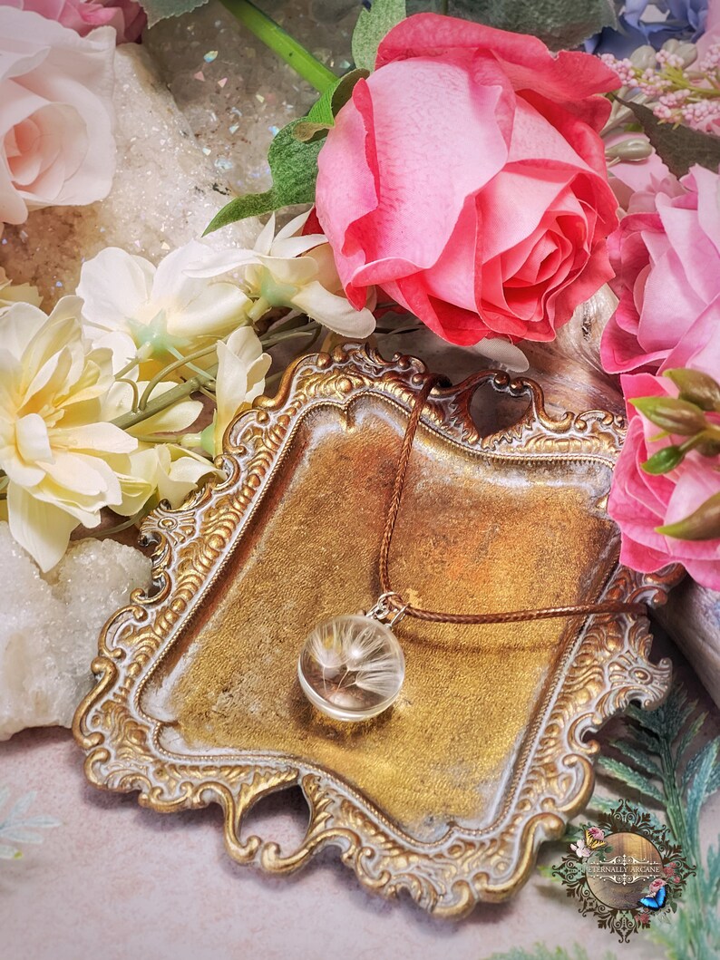 May include: A delicate necklace with a glass pendant containing a dried dandelion. The necklace is displayed on a gold-colored tray with a brown cord.