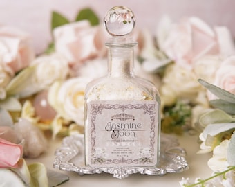 May include: A clear glass bottle filled with white bath salts, topped with a crystal stopper. The label reads "Jasmine Moon Divine Feminine" and is surrounded by floral accents. The bottle sits on a decorative silver tray, with a soft floral background.