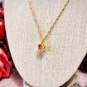 May include: A gold chain necklace with a small red and gold pomegranate pendant. The necklace is displayed on a beige fabric mannequin.