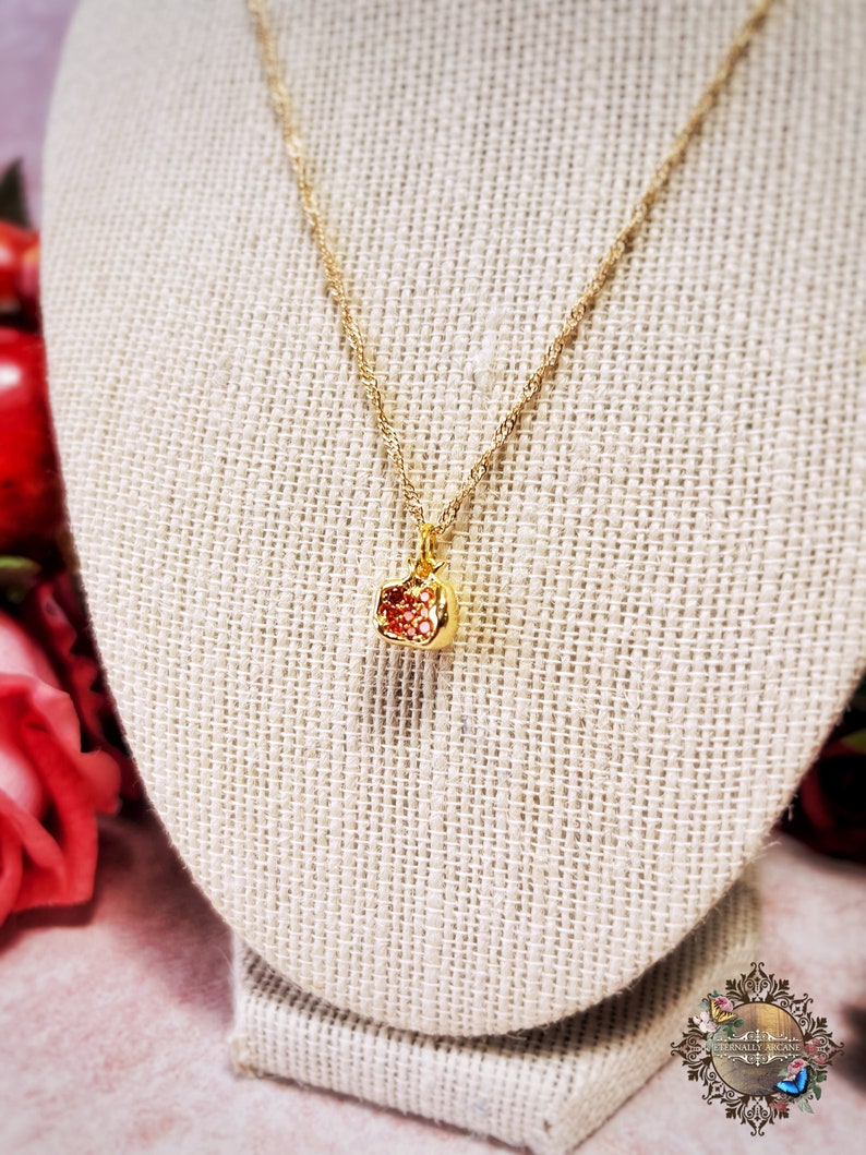 May include: A gold chain necklace with a small red and gold pomegranate pendant. The necklace is displayed on a beige fabric mannequin.