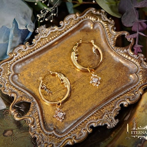 May include: Gold-toned hoop earrings with celestial and gemstone charms rest on a decorative gold tray. The earrings feature a crescent moon and star design, with a dangling, sparkling, star-shaped charm. The tray has ornate edges and is surrounded by flowers.