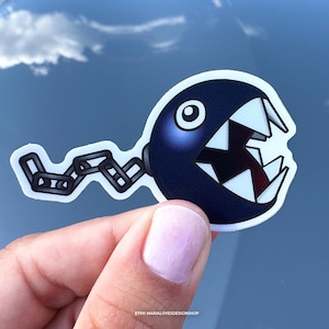 May include: A sticker featuring a cartoon Chain Chomp character from the Super Mario video game series. The Chain Chomp is dark blue with a white mouth, sharp teeth, and a black chain. The background is a blue sky with clouds.