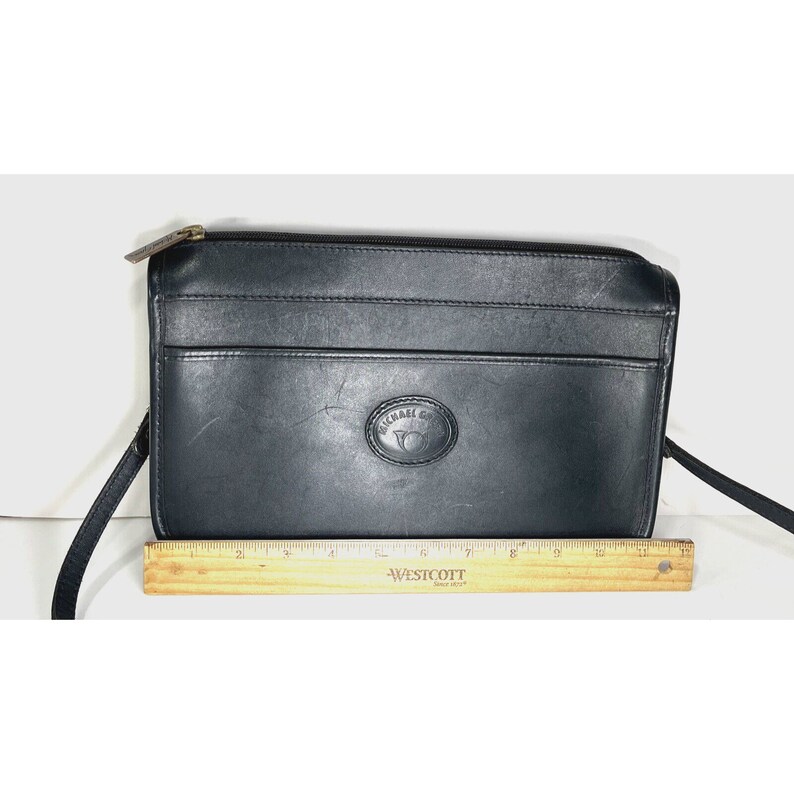 May include: Black leather crossbody bag with a top zipper closure and a front pocket. The bag features a logo on the front and a long strap. The bag measures approximately 12 inches wide.