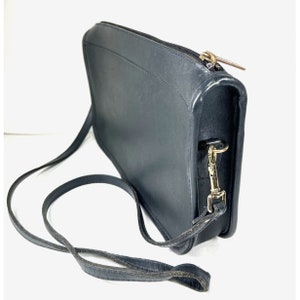 May include: Black leather crossbody bag with a long, adjustable strap. The bag features a rectangular shape with a zipper closure and gold-toned hardware. The leather appears slightly worn, adding to its vintage appeal.