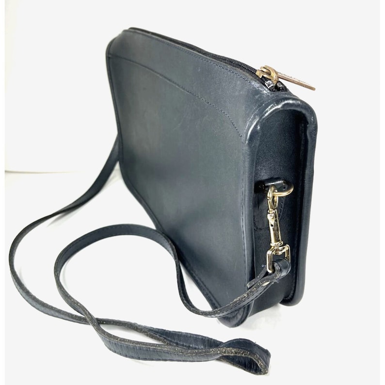 May include: Black leather crossbody bag with a long, adjustable strap. The bag features a rectangular shape with a zipper closure and gold-toned hardware. The leather appears slightly worn, adding to its vintage appeal.