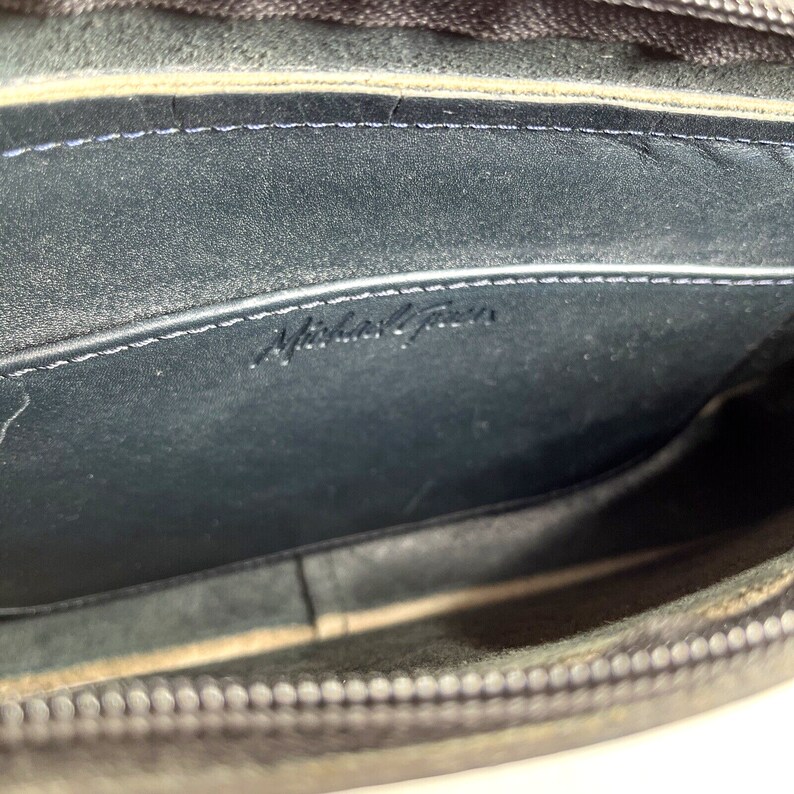May include: Interior view of a black leather handbag. The inside is dark with a stitched seam and the embossed text "Michael Kors". The zipper is visible at the top, and the bag appears to be well-worn.