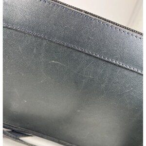 May include: Close-up of a black leather accessory with a zipper. The leather has a textured appearance with visible scratches. The zipper is black and runs along the top edge. The item appears to be a wallet or small bag.