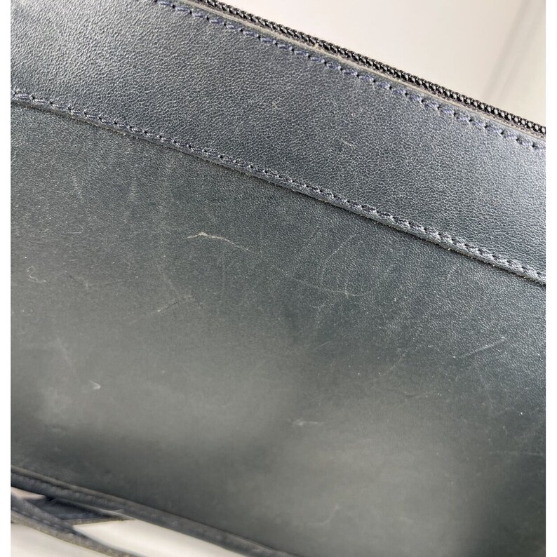 May include: Close-up of a black leather accessory with a zipper. The leather has a textured appearance with visible scratches. The zipper is black and runs along the top edge. The item appears to be a wallet or small bag.