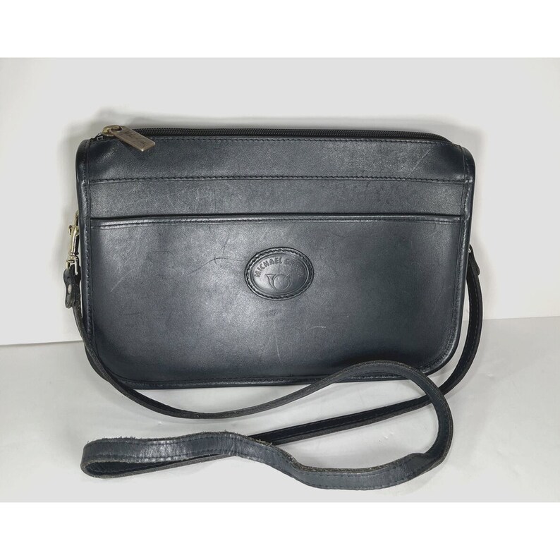 May include: Black leather crossbody bag with a long strap. The bag has a rectangular shape with a top zipper closure and a front pocket. The brand name "MICHAEL G" is embossed on the front.