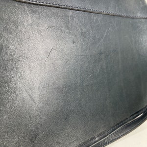 May include: Close-up of a black leather bag, showing the textured surface and stitching details. The bag has a worn appearance with visible scratches and marks, suggesting use. The image highlights the material and craftsmanship of the accessory.