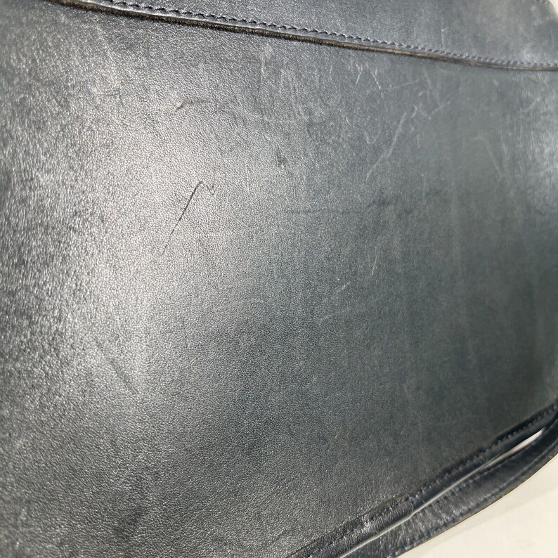 May include: Close-up of a black leather bag, showing the textured surface and stitching details. The bag has a worn appearance with visible scratches and marks, suggesting use. The image highlights the material and craftsmanship of the accessory.