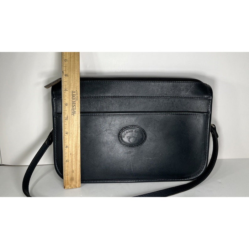 May include: Black leather crossbody bag with a long adjustable strap. The bag has a rectangular shape with a zippered top and a front pocket. The bag measures approximately 9 inches long. The brand name "MICHAEL CO" is embossed on the front.