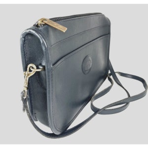 May include: A navy blue leather crossbody bag with a long strap. The bag has a zippered top, a front pocket, and a gold-tone zipper pull. A circular logo is visible on the front of the bag.