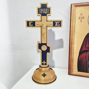 May include: A gold and navy blue Orthodox cross with a figure of Jesus Christ. The cross has inscriptions in an unknown language and sits on a gold base. A religious icon is visible in the background.