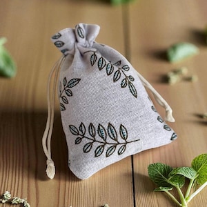 May include: A small, light gray drawstring bag with a botanical leaf pattern in brown and teal. The bag is made of a textured fabric and has a cream-colored drawstring closure. The bag is sitting on a wooden surface.