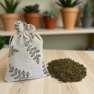 May include: A small, beige drawstring bag with a green leaf pattern, next to a pile of dried green herbs. The bag has a light-colored, woven texture and a natural-colored drawstring. The herbs are finely chopped and appear to be dried leaves.