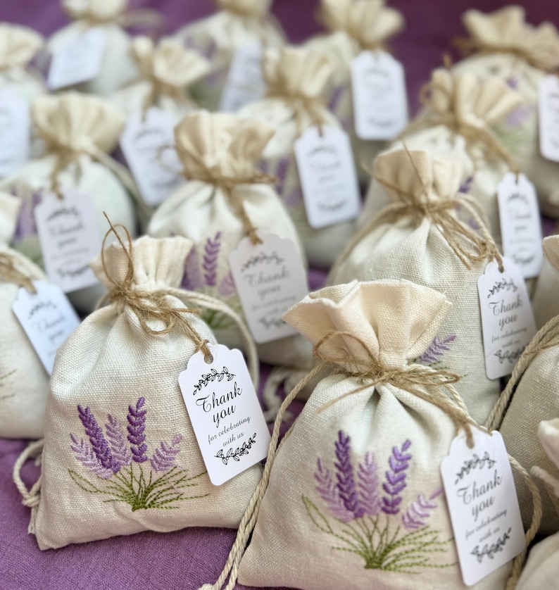 May include: Small, beige linen drawstring bags with embroidered purple lavender sprigs. Each bag is tied with twine and has a white tag that reads "Thank you for celebrating with us."