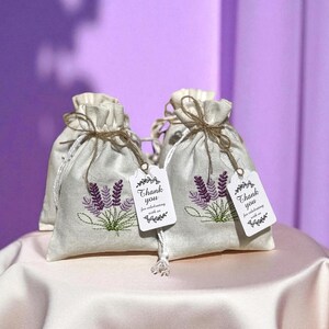 May include: Two beige drawstring bags with embroidered lavender sprigs and attached tags that read "Thank you". The bags are tied with twine and sit on a satin surface, against a purple backdrop.