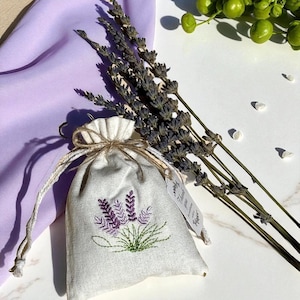 May include: A small, off-white linen sachet with a drawstring closure, embroidered with purple lavender sprigs and green leaves. Dried lavender stems and a purple cloth are arranged nearby. A small tag is attached to the sachet.