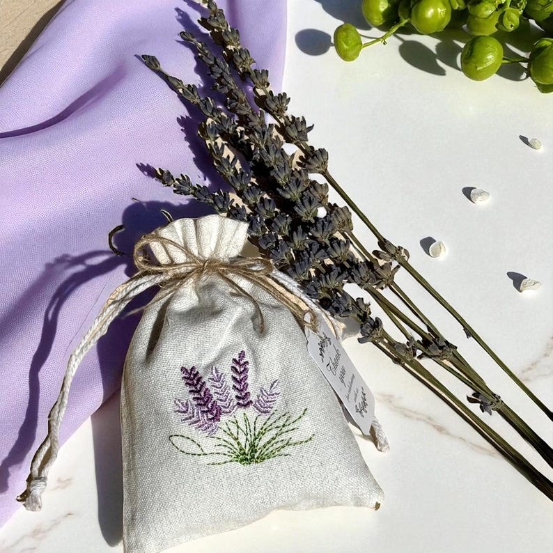 May include: A small, off-white linen sachet with a drawstring closure, embroidered with purple lavender sprigs and green leaves. Dried lavender stems and a purple cloth are arranged nearby. A small tag is attached to the sachet.