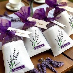May include: White sachets filled with lavender, tied with purple ribbon and tags that say "Thank you." Each sachet has a lavender sprig design and the word "Lavender" in a purple rectangle. Dried lavender sprigs are scattered nearby.