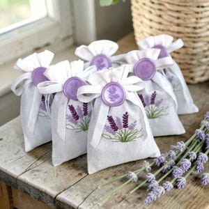 May include: A group of small, light gray fabric sachets, each featuring an embroidered lavender sprig in purple and green. The bags are tied with a white ribbon and sealed with a round, light purple wax seal embossed with a floral design.