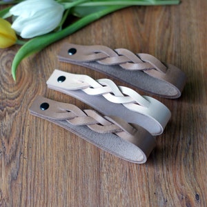 May include: Three braided leather bracelets in brown, beige, and white. The bracelets are on a wooden surface.