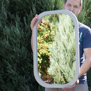 May include: A rectangular mirror with rounded corners, featuring a silver-colored frame. The mirror reflects a green, leafy background with hints of red berries. The frame has a smooth, matte finish.