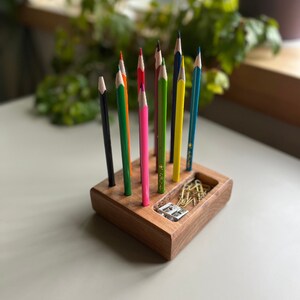 May include: A wooden desk organizer with a variety of colored pencils, a pencil sharpener, and paper clips. The pencils are arranged in a row, with colors including black, green, pink, yellow, and blue. The organizer is made of light brown wood.