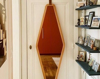 May include: A diamond-shaped wooden mirror with a light brown frame hangs on a white door. The mirror is suspended by a brown leather strap. The reflection shows a red-orange cabinet and wooden floor. A series of black shelves with framed photos are to the right.